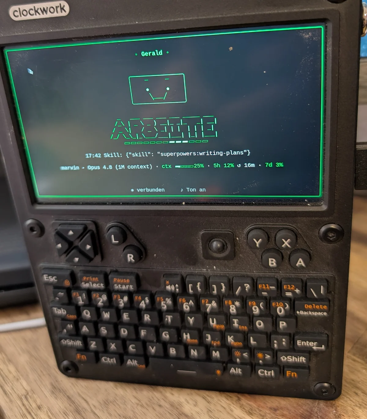 The uConsole handheld on a desk. On its display, in green: the name Gerald, a drawn face with raised brows, below it the large status word “ARBEITE” with a running bar, the current tool call, and a status line showing model, context fill and usage limits.